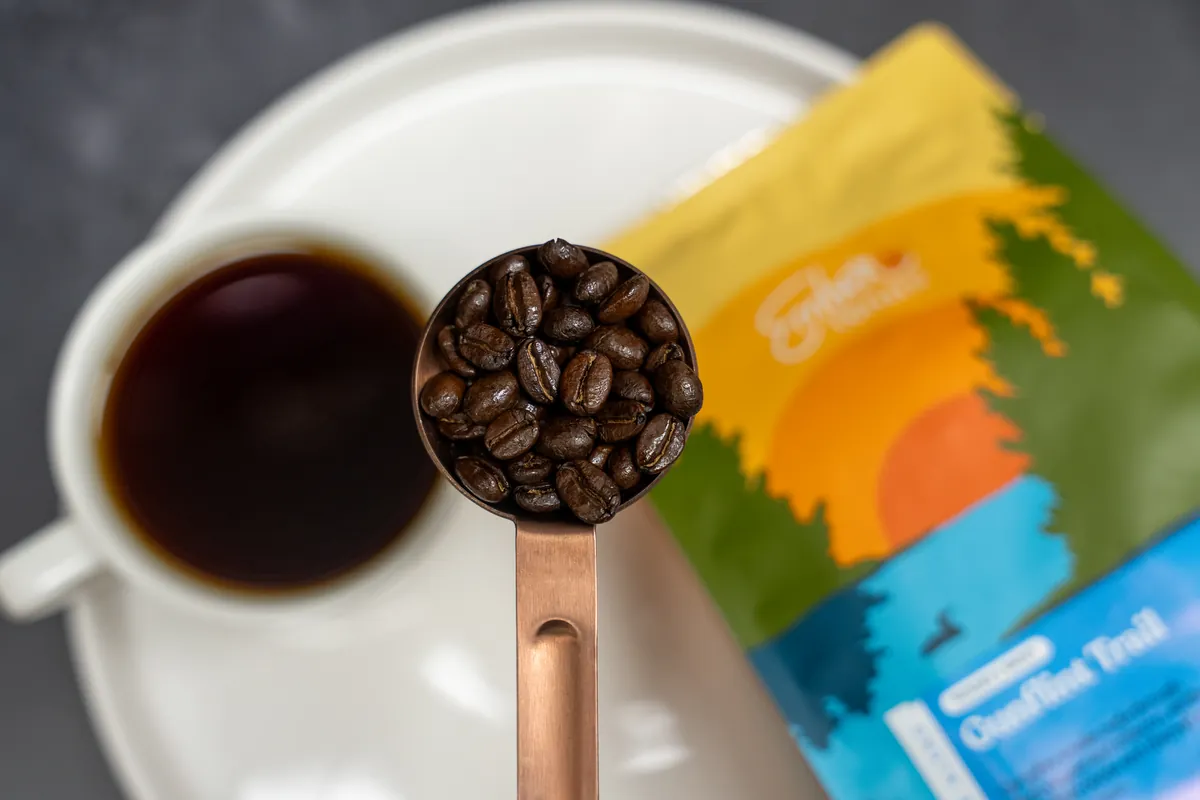 A copper scoop of roasted Ember coffee beans over a cup of black coffee and a bag of Ember coffee
